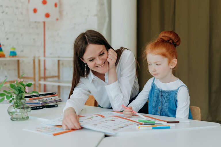 A smiling teacher assists a young girl with a colorful educational activity at a classroom table.
